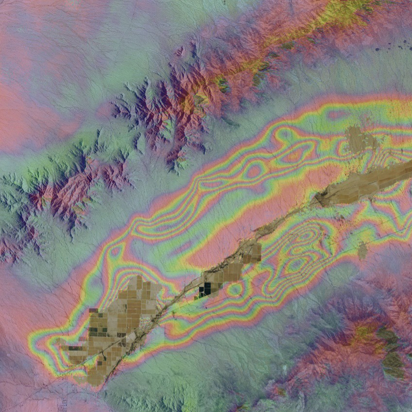 Square image with colors indicating changes in land elevation; yellow/brown area in center of image indicates road and farm fields. Mountains on either side of colored fringes.