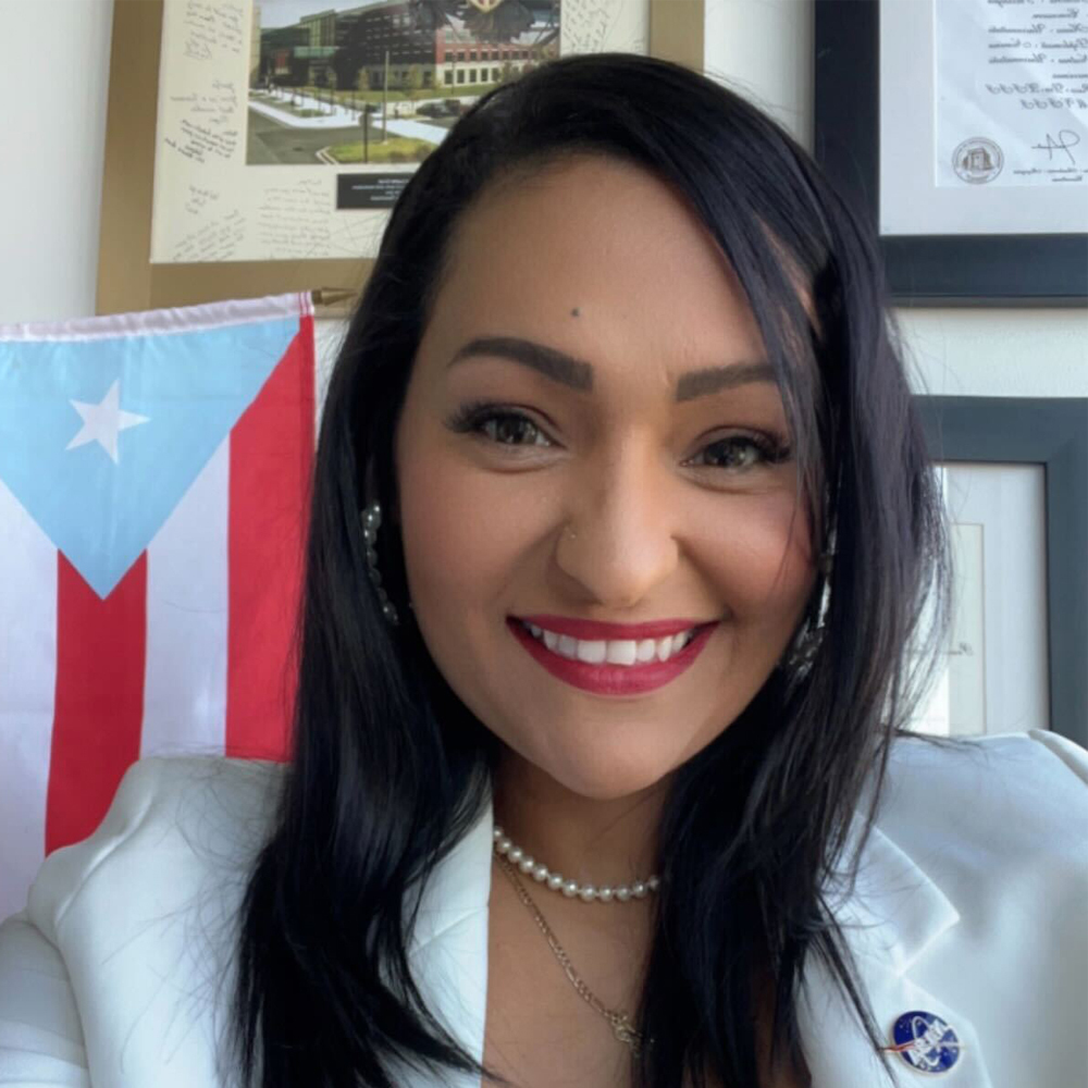 Dr. Yaitza Luna-Cruz smiling and wearing a white dress coat while standing in front of a wall of office plaques and a Puerto Rican flag.