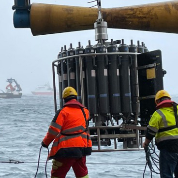 This thumbnail image shows two crewmembers on a ship preparing to lower a scientific instrument called a rosette into the ocean. The two crewmembers are holding ropes and steadying the instrument. They are wearing orange and green ocean gear and hard hats. The rosette is about six feet tall and consists of many long, gray cylinders oriented in a vertical circle and ringed by a strong, metal frame. 