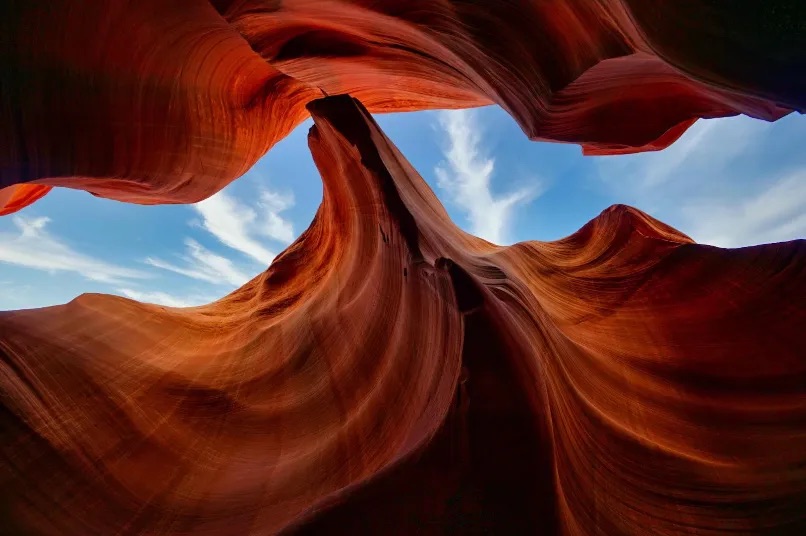 Antelope Canyon photographed from the ground looking up through the red cliffs against a blue sky streaked with clouds