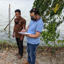 Two citizen scientists standing on the shore of a lake collecting water height data from a gauge in the background.