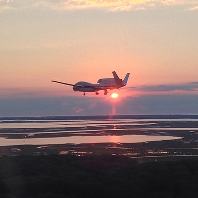 This image shows a Delta Hawk drone as seen from the window of a chase plane. The drone is positioned in the middle of the frame and flying over a marine marsh area at sunset. The Sun is just below the bottom of the drone and about to disappear behind a cloudbank on the horizon. 
