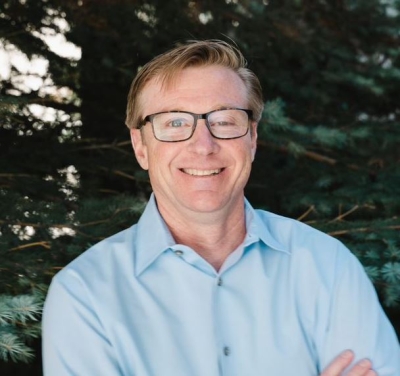 A headshot of Dr. Eric Wilcox, Research Professor of Atmospheric Science at the Desert Research Institute. Dr. Wilcox, who is wearing glasses and a light blue oxford shirt is standing, arms crossed, before a tree-filled background. 