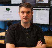 Dr. Dan Runfola, Associate Professor of Applied Science, William & Mary, sits arms crossed at a desk in front of several computer monitors in a lab.