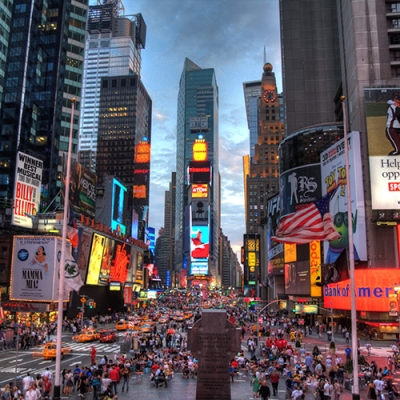 This image features of view New York City's Times Square looking south in early evening. The right, left, and middle background of the image shows office buildings, skyscapers, and stores with bright lights and advertising signs surrounding the square. The center of the image shows cars and people traversing the streets and sidewalks below.