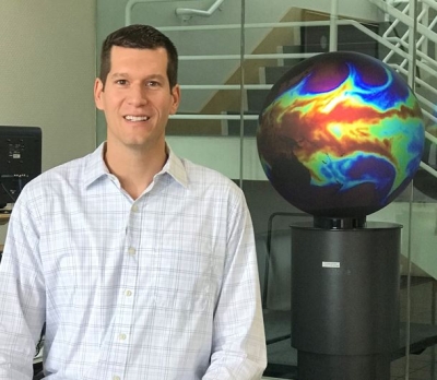 Dr. David Peterson, Naval Research Laboratory meteorologist poses with a globe showing wavy patterns of satellite data. Peterson is in a white, button-down shirt and sitting in the lobby of a large office building. A staircase with a white railing can be seen in the background.