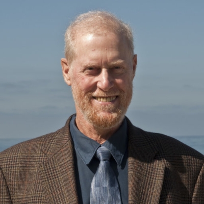 A headshot of Dr. Yehuda Bock, Distinguished Researcher and Senior Lecturer at the Scripps Institution of Oceanography’s Institute of Geophysics and Planetary Physics. Dr. Bock is standing before an ocean backdrop, wearing a brown suitcoat, dark gray shirt, and gray tie.