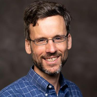 headshot of adam storeygard wearing a blue check shirt and standing in front of a brown screen