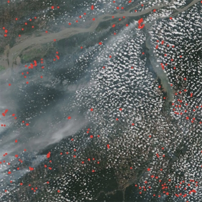 This is a satellite view of wildfires across a portion of the Amazon Rainforest. The image shows popcorn clouds and wispy streaks of fire smoke over the forest and winding Amazon River. Overlayed across the image are hundreds of small red dots to mark the location of fires identified by satellite sensors.