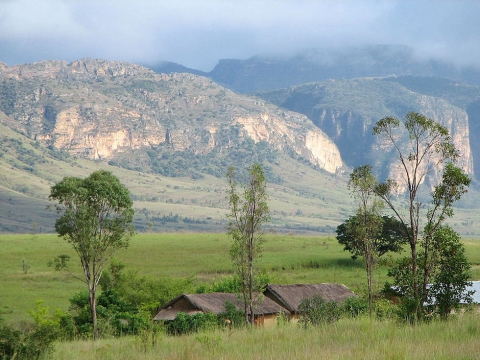 This image is of Isalo National Park in Madagascar. The foreground of the image shows a tall grassy field. In the middle of the field are a few trees and three small rural-style buildings or barns. In the background are low mountains with dark clouds shrouding the peaks..