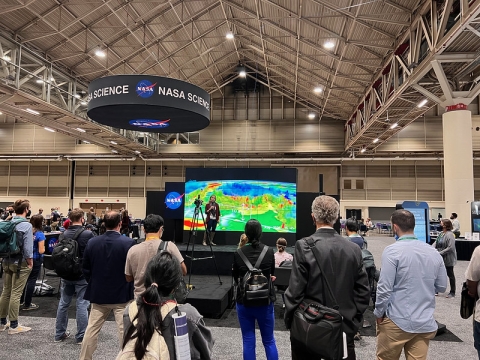 Image of NASA Hyperwall presentation with people watching; NASA Science banner hangs from ceiling.