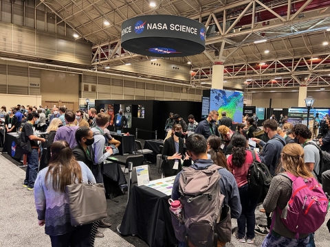 Conference attendees milling around tables at the NASA Booth; NASA Science banner hangs from ceiling.