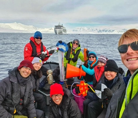 Outdoor image of 9 scientists in a motorboat with icebergs behind them.