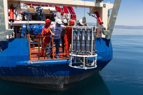 This image shows five technicians wearing hard hats and overalls lowering a device called a rosette into the water off the back of a blue research vessel on a calm, sunny day.. The rosette is a device roughly six feet tall made up of a cluster of gray cylinders arranged in a circle secured inside a white frame. 