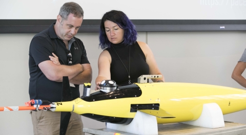 Dr. Jeremy Werdell and Dr. Ivona Cetinić stand in a well-lit room with a yellow ocean glider in front of them. Werdell is wearing a black golf shirt and Cetinić is wearing a sleeveless black shirt. The glider is about four feet long and resting on a table. The glider has a streamlined shaped, with an orange rod projecting from its nose, a short round device on its top, and short wings extending from its side.