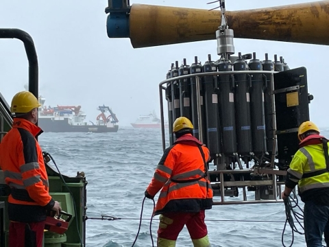 This image shows three crewmembers on a ship preparing to lower a scientific instrument called a rosette into the ocean. Two crewmembers are holding ropes and steadying the instrument. All three people are wearing orange and green ocean gear and hard hats. The rosette is about six feet tall and consists of many long, gray cylinders oriented in a vertical circle and ringed by a strong, metal frame. In the background are two other ships floating in mildly a wavy ocean with an overcast sky.