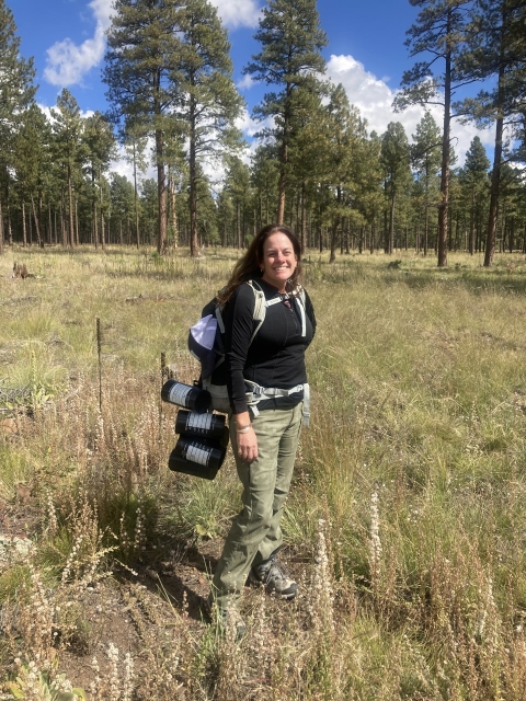 Dr. Helen Poulos stands in a meadow on the edge of a pine forest in the southwestern United States.