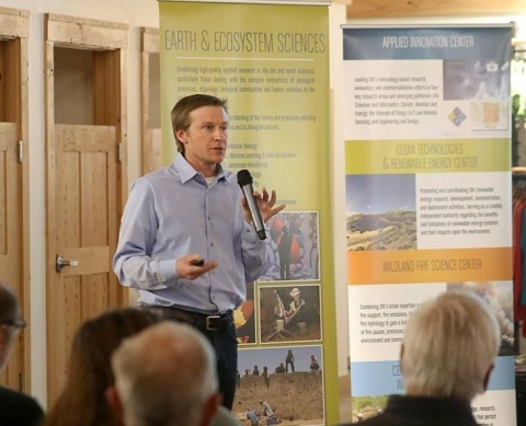 Dr. Eric Wilcox, in a blue shirt and standing in front of displays about Earth and ecosystems sciences research, speaks to an audience at a public Desert Research Institute event in Nevada.