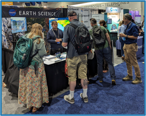 Image of NASA booth at Esri with numerous people standing in front and one NASA representative inside the booth interacting with visitors