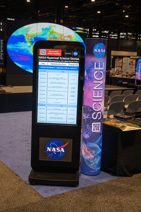 Electronic schedule at entrance to NASA Booth with NASA Science illuminated pillar to right of schedule