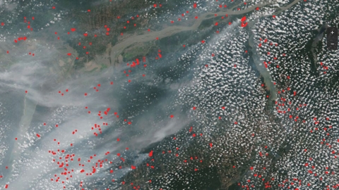 This is a satellite view of wildfires across a portion of the Amazon Rainforest. The image shows popcorn clouds and wispy streaks of fire smoke over the rainforest and winding Amazon River. Overlayed across the image are hundreds of small red dots to mark the location of fires identified by satellite sensors.