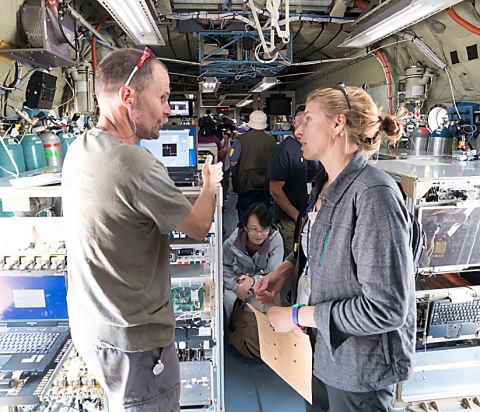 Image of Dr. Fischer talking with her colleague Dr. Frank Flocke inside a C-130 research aircraft. The aircraft interior is bare steel with instruments throughout the cabin. Fluorescent lights hang from the ceiling.