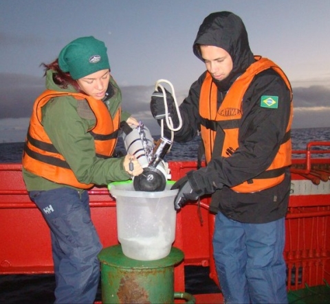 Image of Dr. Lange and a colleague standanding aboard a ship in the open ocean and wearing life vests and other protective gear. Both scientists are dumping water from a collection bottle into a sampling container. Dr. Araujo wears a Brazilian flag patch on his jacket and stands on Dr. Lange's left side.