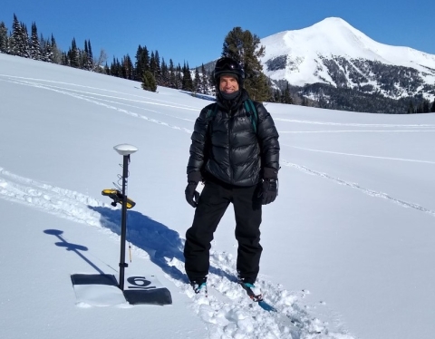 Dr. Sproles wearing all black snowgear standing on fresh white snow next to a GPS receiver. A mountain in the background has fresh snow on top, grading to bare black earth farther down the slope.