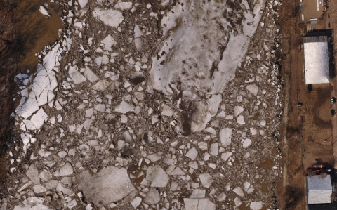 Overhead image of whitish blocks of ice on a wide point of the Yellowstone River. On the right side bank are two rectangular sheds along with several cars and people.