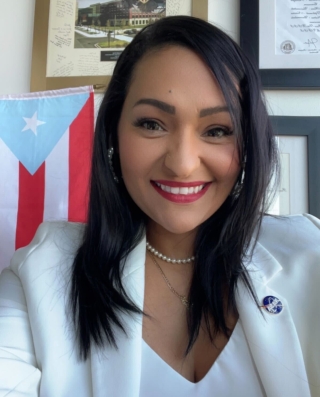 Dr. Yaitza Luna-Cruz smiling while wearing a white dress jacket while standing in front of a wall of plagues and a Puerto Rican flag.