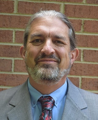 Dr. Gerald (Stinger) Guala wearing a blue shirt, a necktie, and gray suit standing in front of a red brick wall.