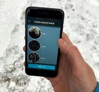 A picture of a hand holding a smartphone over snow-covered ground. The smartphone shows an application open to select which of three types of precipitation are currently being observed.