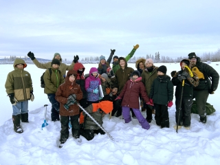 A group of students wearing winter clothing pose for a picture on a snow-covered river. There are clouds and a forest in the background.