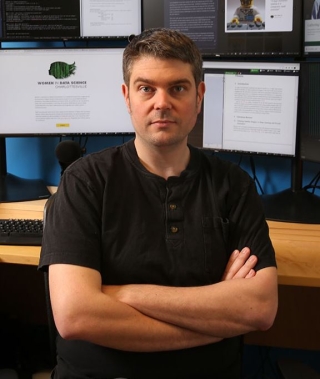Dr. Dan Runfola, Associate Professor of Applied Science, William & Mary and member of SEDAC's User Working Group, sits arms crossed at a desk in front of several computer monitors in a lab.
