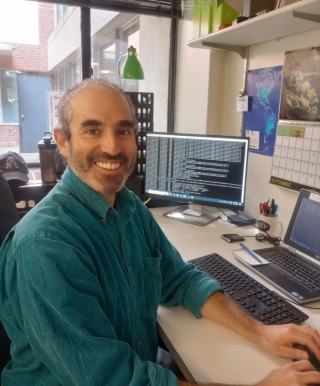 Dr. Gonzalo González Abad, an Atmospheric Physicist at the Center for Astrophysics, sits at a desk in his office. He is wearing a green collared shirt and behind him is a computer monitor showing code used to manipulate data.