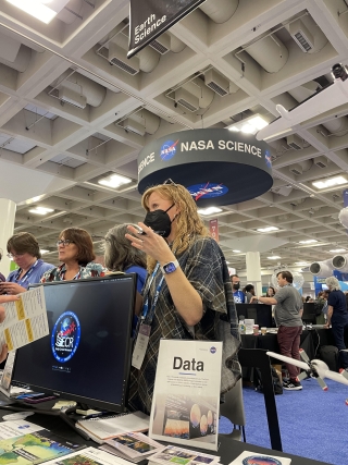 Image of woman wearing a mask talking with a guest at a table with a sign reading Data on it; banner saying NASA Science is above her