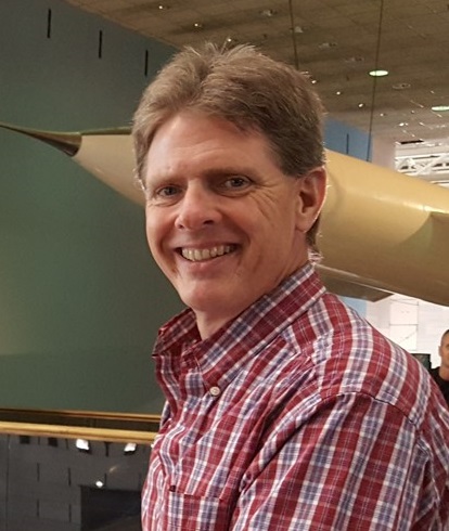 Headshot of Dr. Kurt Thome wearing a red plaid Oxford shirt and standing in front of a rocket suspended from the ceiling of the National Air and Space Museum.
