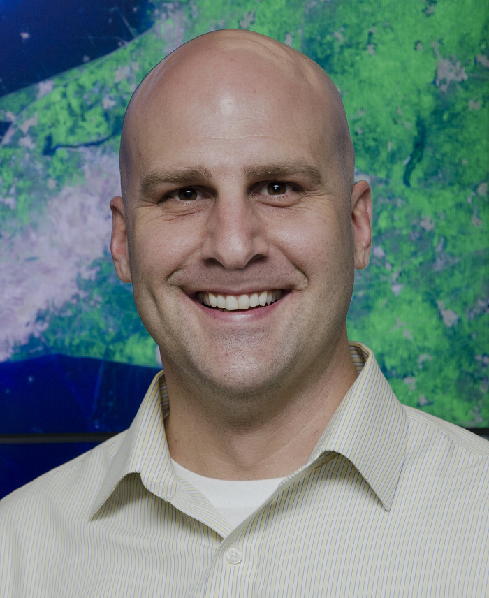 Headshot of Mark McInerney wearing an open collar Oxford shirt and standing in front of a map.