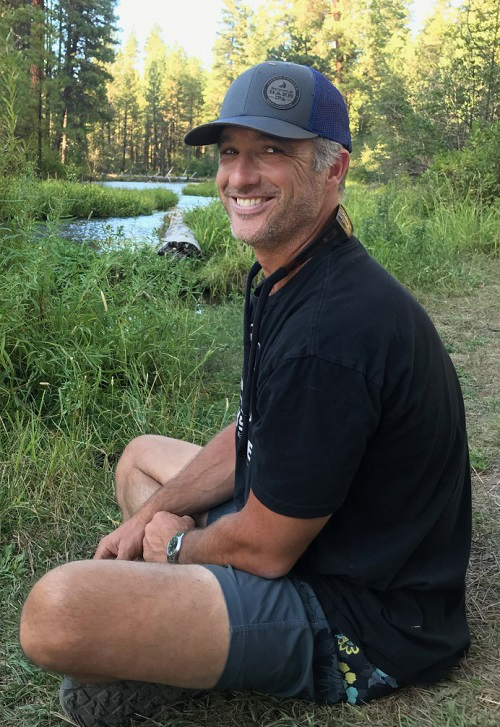 Image of Dr. Eric Sproles sitting by the side of a stream in a wooded area wearing shorts, a T-shirt, and a baseball cap.